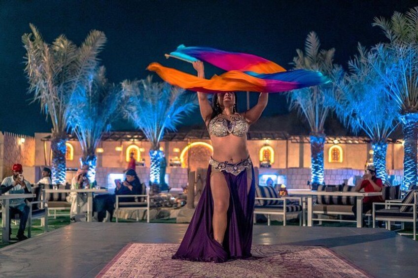 Een buikdanseres treedt op met kleurrijke sjaals voor een verlicht gebouw met palmbomen. 1