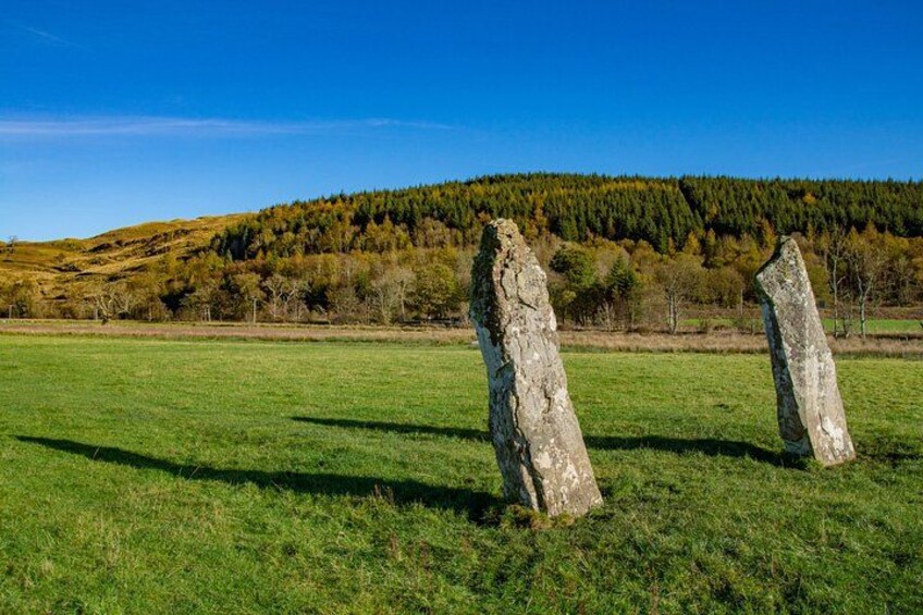 Two standing stones in a grassy field with a forested hill in the background. 1