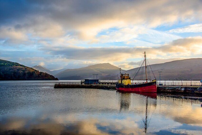 A calm lake with a red boat docked at a pier, surrounded by mountains and a cloudy sky. 2