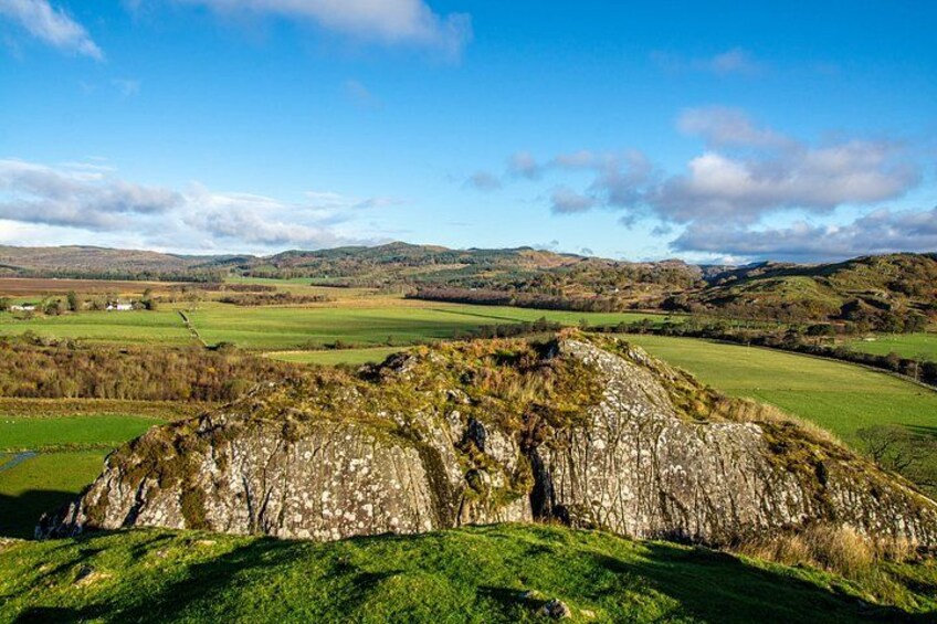 A rocky hill with a grassy foreground, rolling hills, and a clear blue sky in the background. 3