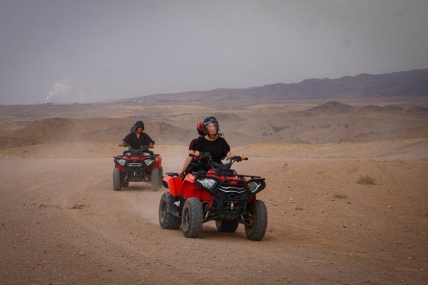 Two people riding ATVs on a dusty road with a desert landscape in the background. 1