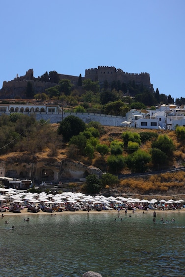 A coastal town with a historic castle on a hill, a beach with umbrellas, and people swimming in the water. 3