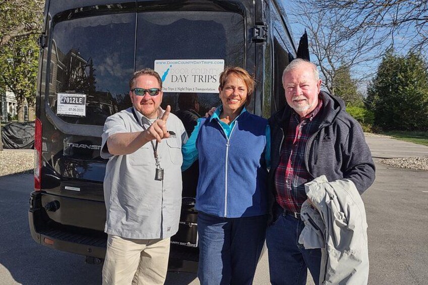 Three people standing in front of a black bus with a sign that reads 'Door County Day Trips.' 2