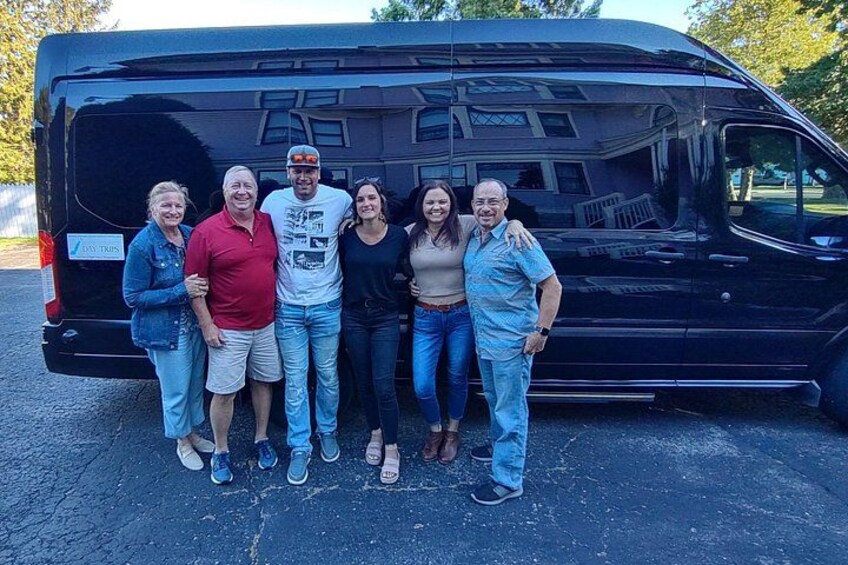 A group of people standing in front of a black van with a house reflected in the rear window. 5