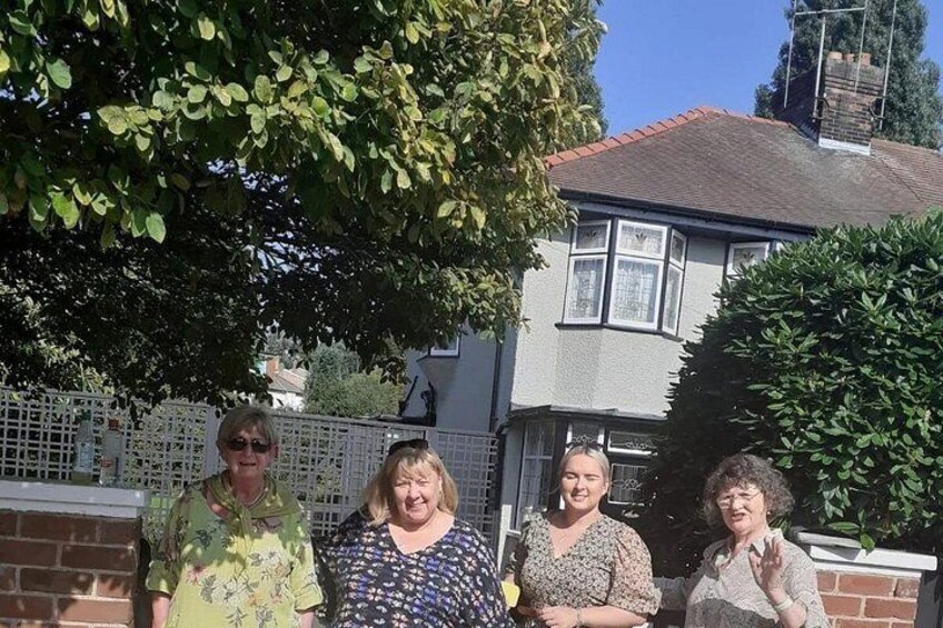 Cuatro personas de pie frente a una casa con un muro de ladrillo y un árbol. 5
