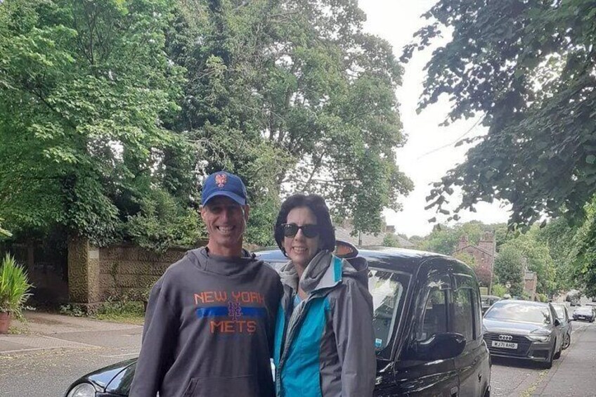 Dos personas de pie frente a un coche negro en una calle con árboles y una valla al fondo. 3