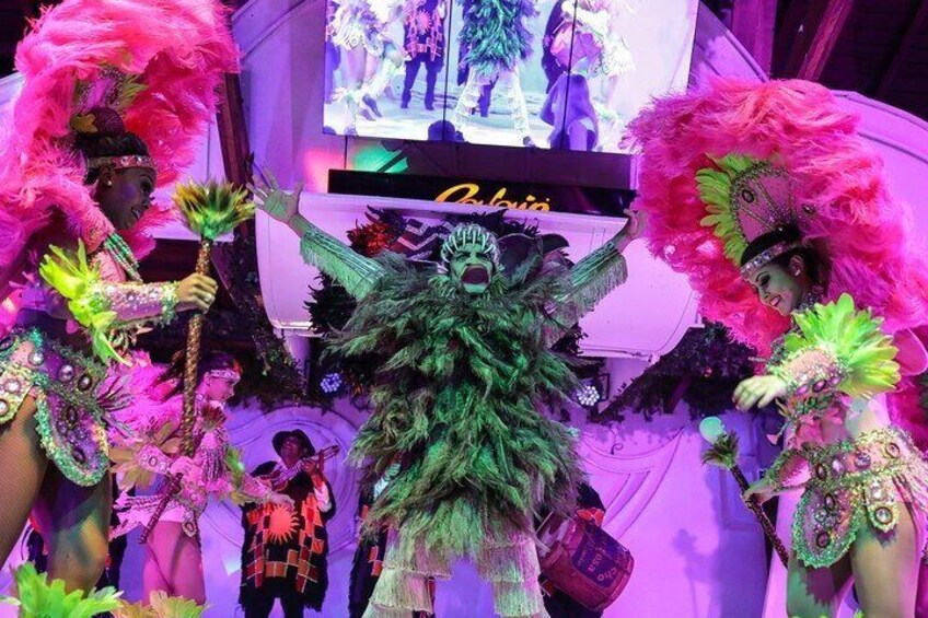 A person in a green feathered costume with a large headdress, surrounded by others in vibrant costumes. 1