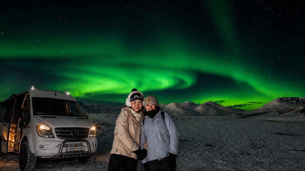 Two people standing next to a white vehicle under a night sky with green aurora. 1