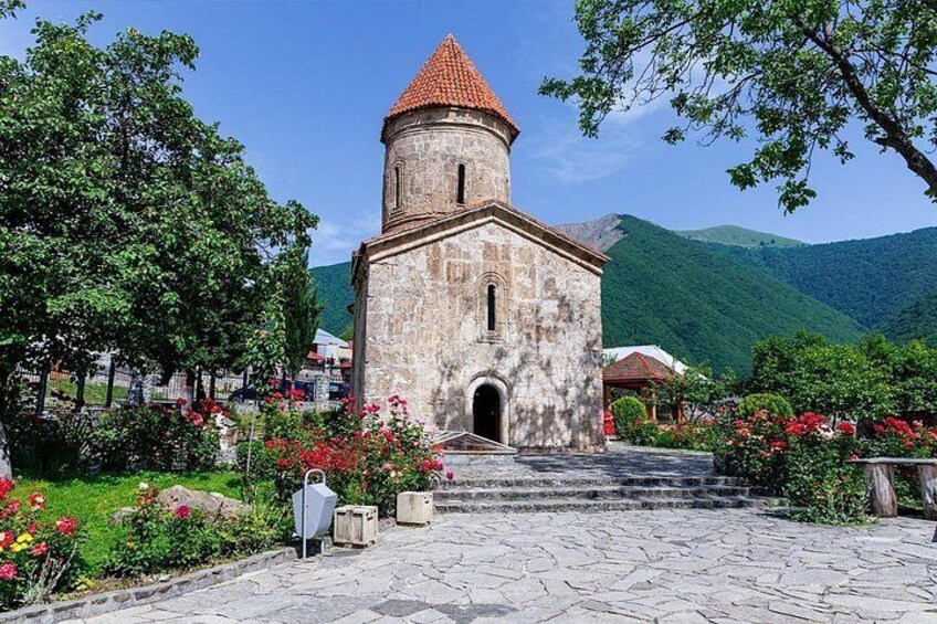 A stone church with a red-tiled roof, surrounded by lush greenery and a mountainous backdrop. 3