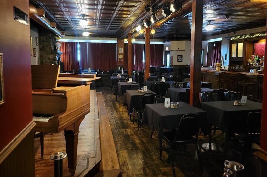 A restaurant interior with wooden tables, chairs, a piano, and a bar area. 3