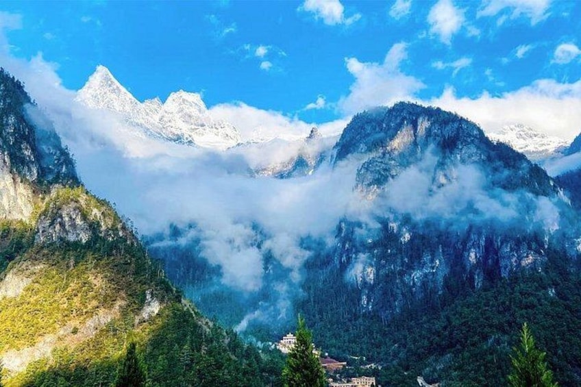 A mountainous landscape with snow-capped peaks, dense forests, and a small building at the base. 1