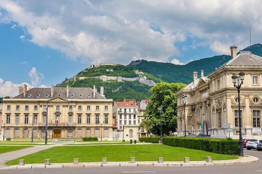 A historic building complex with a green lawn in front, a hill with a fortification in the background, and a clear sky. 5