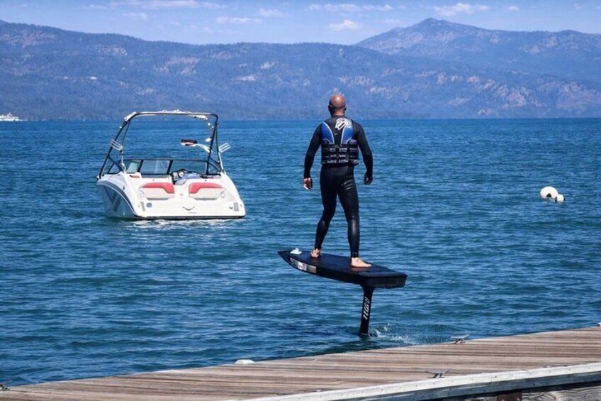 Una persona de pie en una tabla de remo en el agua, con un barco al fondo y montañas a lo lejos. 3