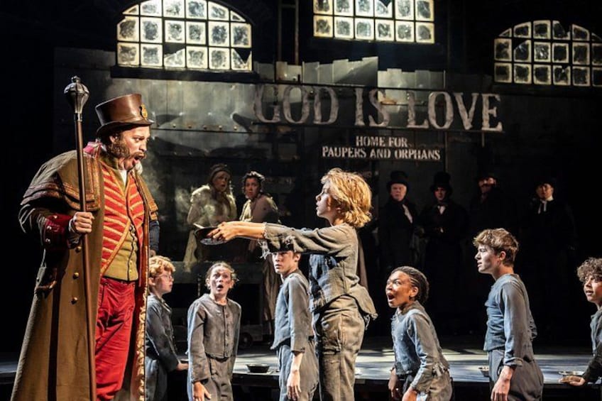 Un palcoscenico teatrale con attori in abiti d'epoca, un cartello con la scritta 'GOOD IS LOVE' e uno sfondo decorato con quadri. 4