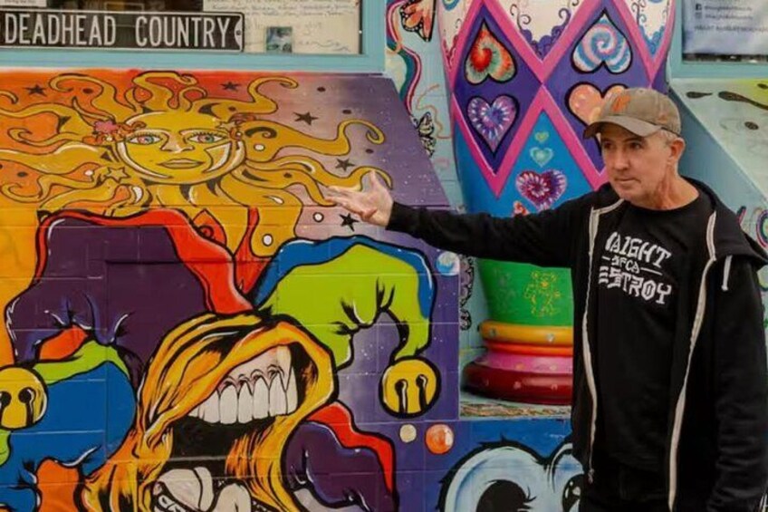 Un mural colorido con una figura central, un hombre señalando, y un letrero que dice 'DEADHEAD COUNTRY'. 1