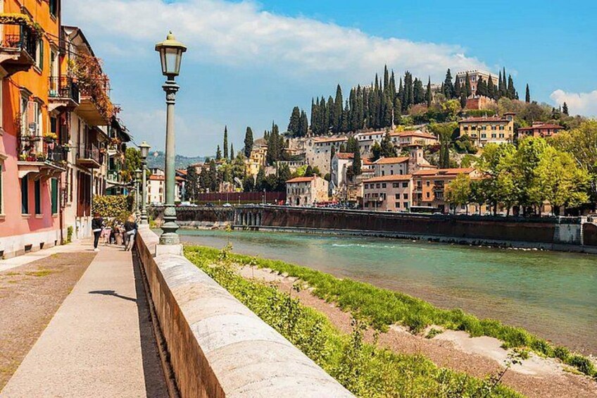A riverside walkway with buildings, a lamppost, and a hill with a castle in the background. 1