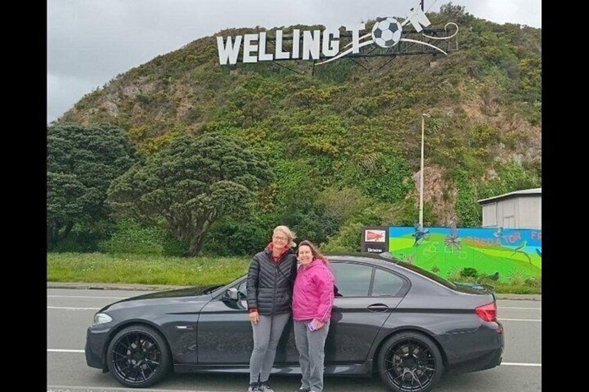 A black car parked on a road with a sign reading 'WELLING' in the background. 4