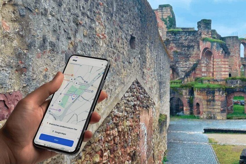 A hand holding a smartphone with a map application open, displaying a location with a green area and a blue button. In the background, there is an old stone structure with arches and a paved path. 1