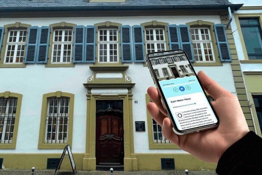 Une main tenant un smartphone qui affiche une carte. Sur la carte, on peut voir un bâtiment portant l’inscription « Kari-Mori-Haus », situé devant un bâtiment bleu clair avec une porte et des fenêtres à volets marron foncé. 5