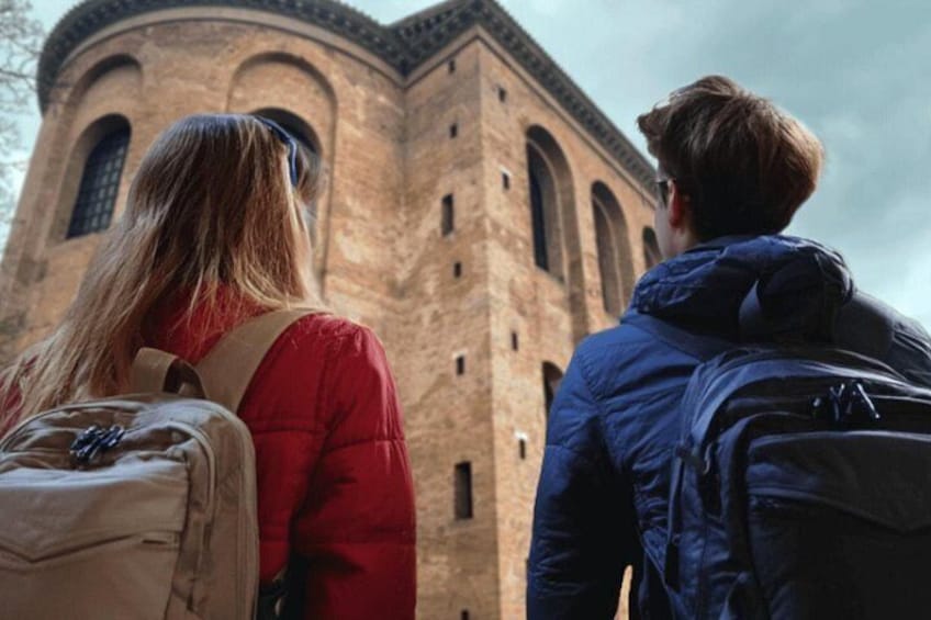 Deux personnes, portant des sacs à dos, se tiennent face à un bâtiment historique en briques, doté d’arches au niveau des fenêtres. 4