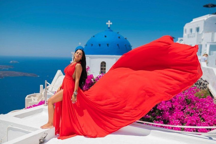 Eine Frau in einem roten Kleid vor einem weißen Gebäude mit einer blauen Kuppel, umgeben von Blumen und dem Meer. 2