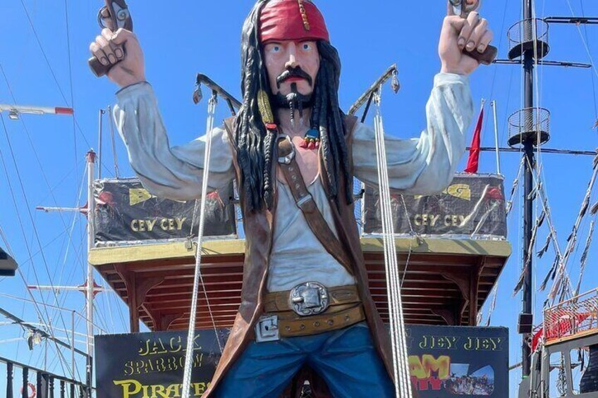 Eine Piratenstatue mit roter Bandana, die ein Schwert und eine Pistole hält und auf einem Schiffdeck mit Segeln und Tauwerk steht. 1