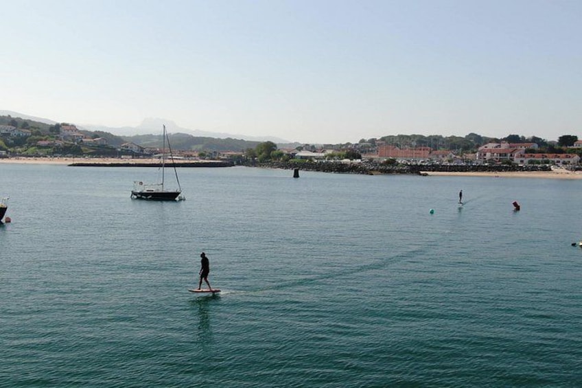 Une mer calme avec un voilier, une personne pratiquant le paddle, et une côte lointaine parsemée de bâtiments. 4