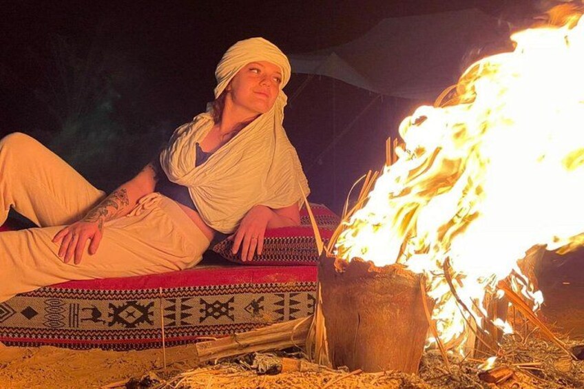 A person sitting on a patterned cushion by a campfire. 4