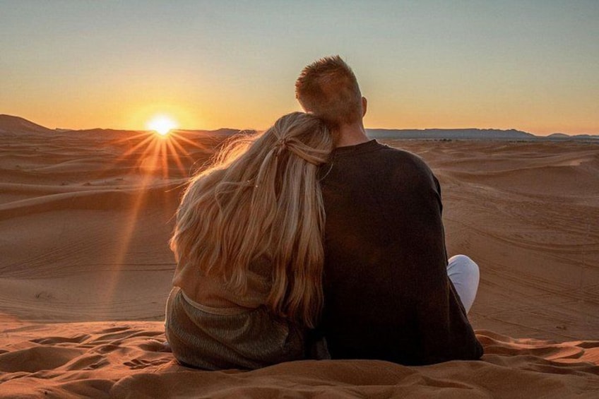 Two people sitting on sand dunes with the sun setting in the background. 1