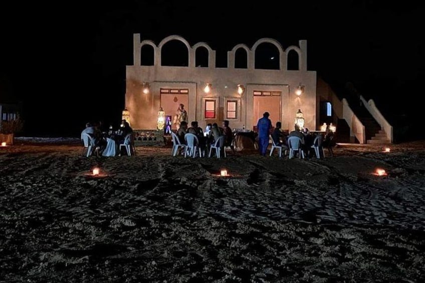 A building with arched windows and a staircase leading up to it, surrounded by people seated at tables with candles on the ground. 5