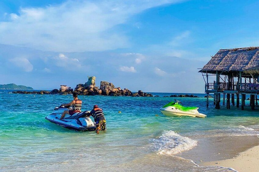 A beach with two jet skis, a thatched-roof structure, and rocky outcrops in the background. 2