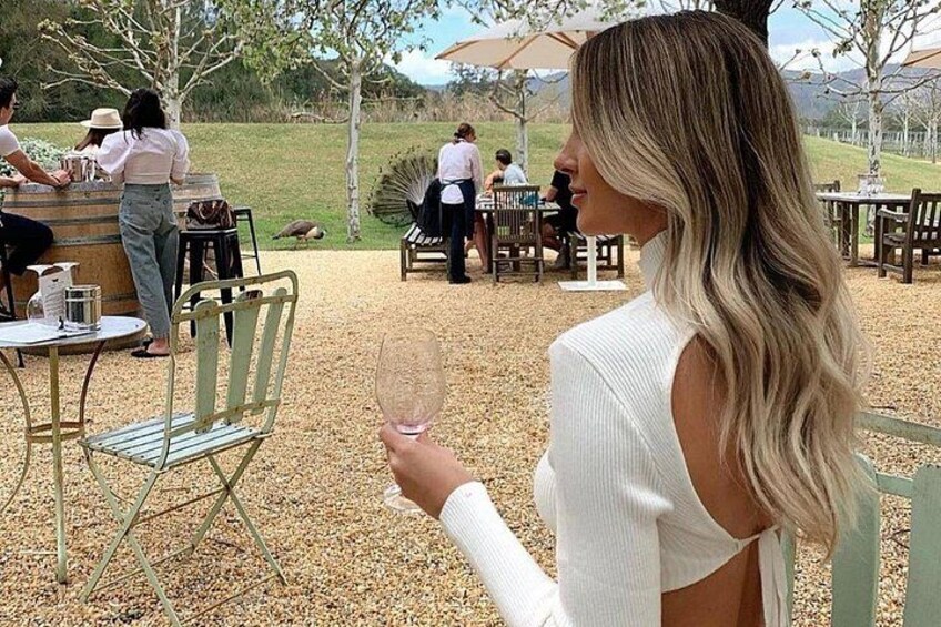 A woman holding a wine glass, seated at an outdoor table with a grassy field and trees in the background. 5