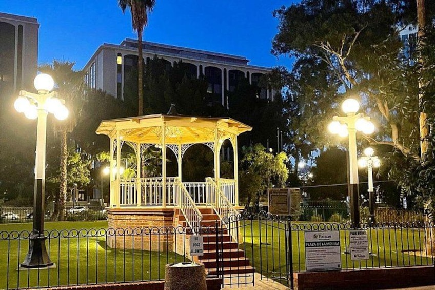 A gazebo with a white railing, surrounded by a fenced area with a grassy lawn, and street lamps. 2