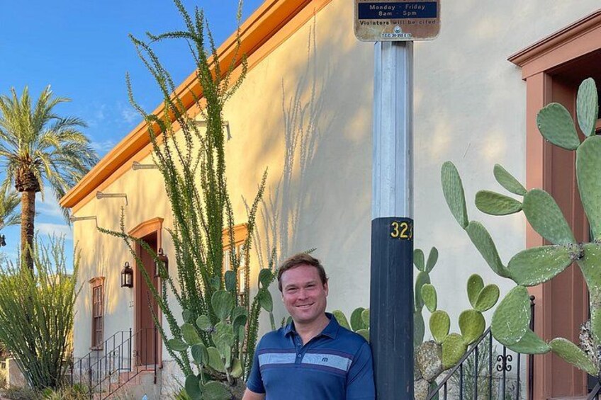 A man standing in front of a building with a signpost, cacti, and a palm tree. 4