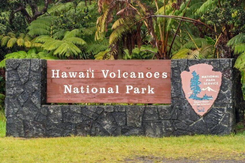 Un panneau indiquant l’entrée du parc national des volcans d’Hawaï, avec un mur de pierres et une végétation luxuriante en arrière-plan. 3
