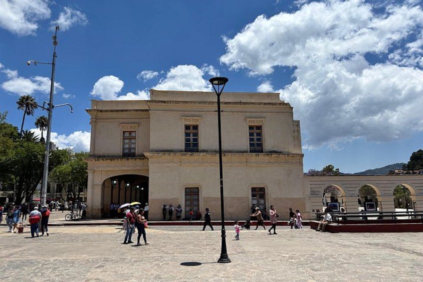Un edificio histórico con ventanas arqueadas y una entrada central, rodeado por una plaza con gente y una farola. 3