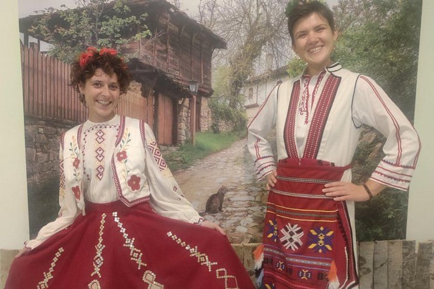 Two people in traditional attire, a red skirt with embroidery, and a white blouse with red and white patterns, standing in front of a rustic wooden house. 4