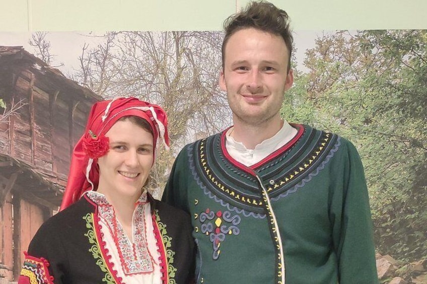 Two people in traditional attire standing in front of a backdrop with a wooden structure and trees. 3