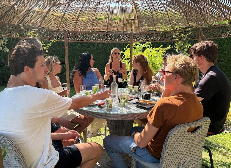 A group of people sitting around a round table outdoors under a thatched roof, engaged in conversation and enjoying drinks. 1