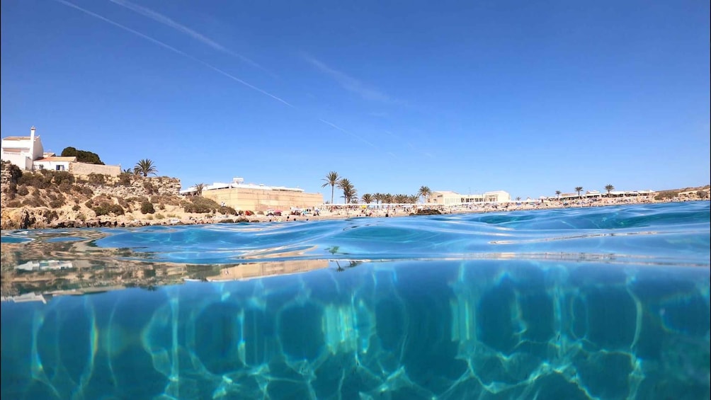 A coastal scene with clear blue water, a sandy beach, and buildings on a hillside. 4
