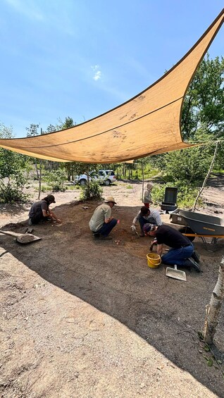 Four people working outdoors under a shade cloth, with a wheelbarrow and tools on the ground. 2