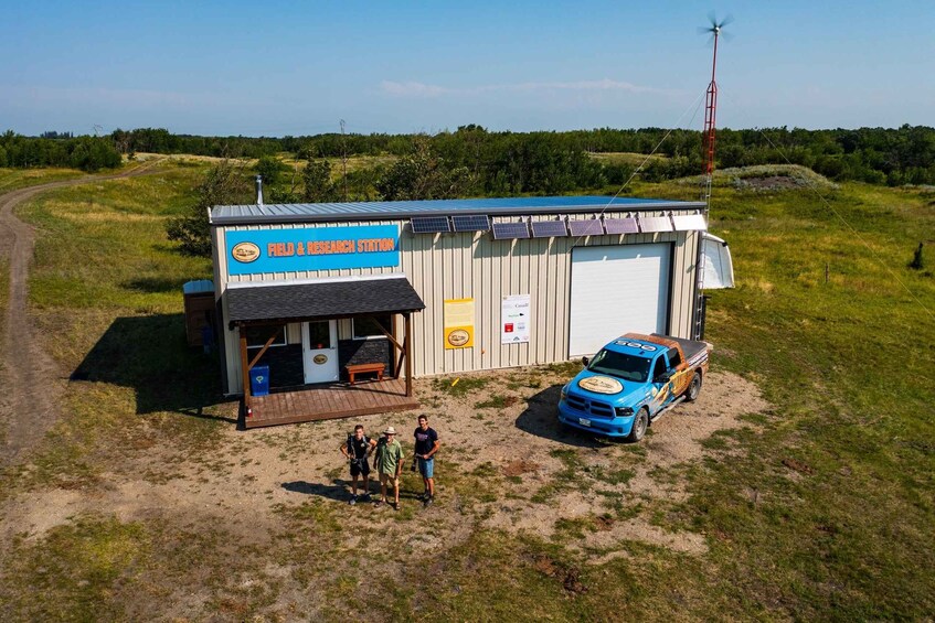 A field research station with a blue building, a parked blue truck, and a wind turbine in a grassy field. 1