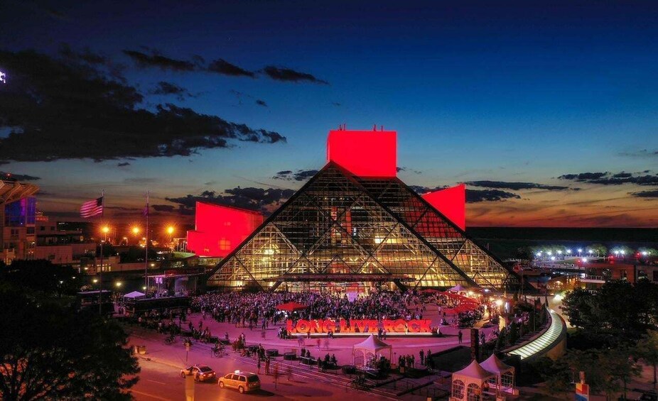 A large, illuminated stadium at dusk with a crowd gathered in front. 1