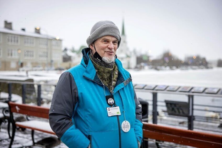 A person standing by a waterfront with snow on the ground, a bench, and buildings in the background. 2
