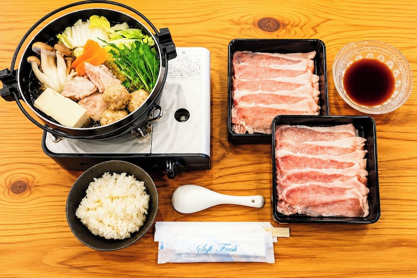 A hot pot with various ingredients, a bowl of rice, a packet of soy sauce, and two trays of raw meat. 3