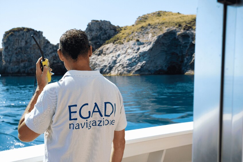 A person on a boat holding a walkie-talkie, with a rocky coastline and clear blue water in the background. 4
