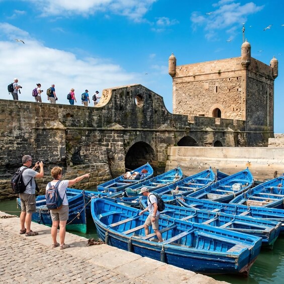 A historic stone tower with tourists on a bridge, and several blue boats docked at the pier. 1