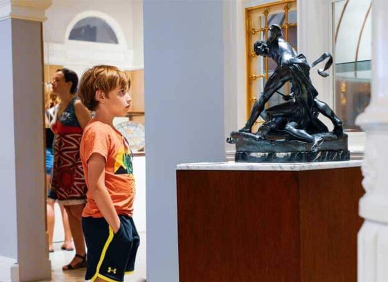 A boy in an orange shirt and black shorts looking at a sculpture of a man in a dynamic pose. 2