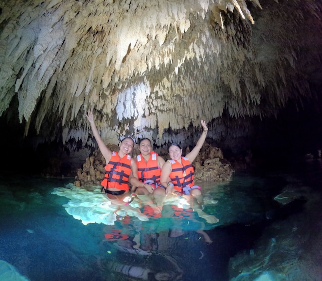 Three people in a cave with stalactites, wearing life jackets and sitting in clear water. 5