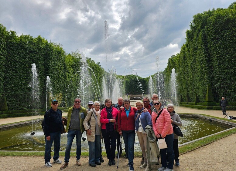 A group of people standing in front of a fountain with water jets in a park with neatly trimmed hedges and a cloudy sky. 5